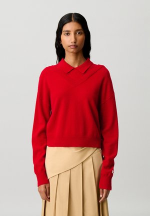 Donna con capelli lunghi e scuri che indossa un maglione rosso brillante con collo e una gonna beige plissettata su uno sfondo chiaro e uniforme.