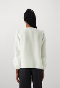 Donna con capelli lunghi e intrecciati, che indossa una camicia bianca a maniche lunghe e pantaloni scuri, vista da dietro su uno sfondo neutro.