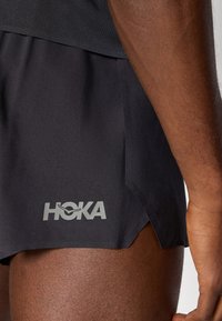 Sorte treningsshorts med slisser på sidene, som har en reflekterende grå "HOKA"-logo. Glatt stoffstruktur med lett design.