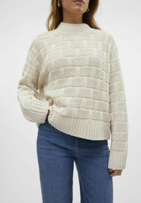 Off-white stickad tröja med ett strukturerat, vävt mönster. Hög krage, långa ärmar, ribbad fåll och lös passform. Kombinerad med blå jeans.