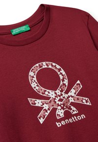 Tmavě červená košile s květinovým vzorem bílého a růžového loga Benetton a zelenou etiketou uvnitř límce.
