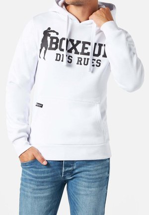 Uomo che indossa una felpa con cappuccio bianca con grafica nera di pugilato e testo "BOXEUR DES RUES", una mano che sistema il cappuccio, l'altra mano in tasca, jeans blu visibili.