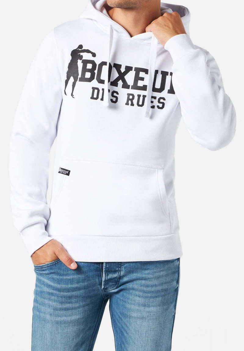 Uomo che indossa una felpa con cappuccio bianca con grafica nera di pugilato e testo "BOXEUR DES RUES", una mano che sistema il cappuccio, l'altra mano in tasca, jeans blu visibili.