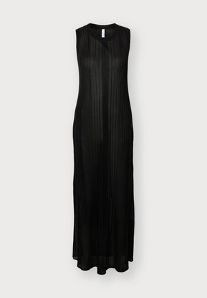 Femme aux longs cheveux blonds portant une robe maxi noire transparente sans manches et des chaussures à talons noires ouvertes à l'orteil, debout devant un fond clair uni.