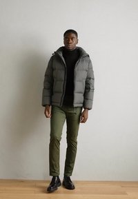 Pufferjack in donkergrijs, met een capuchon en een gestikte ontwerp. Een zwarte coltrui, olijfgroene slim-fit broek en zwarte schoenen maken de look compleet.
