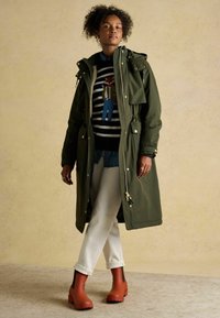 Oliwkowa parka z pełnym zamkiem, regulowanym pasem i dużymi przednimi kieszeniami. Sweter w czarno-białe paski z kolorową haftą. Białe dżinsy i brązowe botki za kostkę.