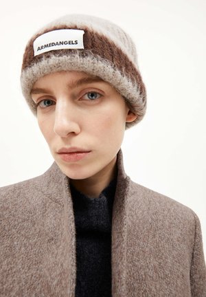 Persoon met een bruin en beige gebreide beanie met het label "ARMEDANGELS", een bruin hoogsluitend jasje en een donkere trui eronder, neutrale achtergrond.