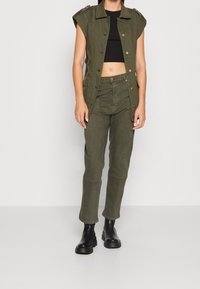 Veste utilitaire sans manches de couleur vert olive avec des boutons-pression et deux poches, associée à un pantalon vert olive assorti et des bottines noires.