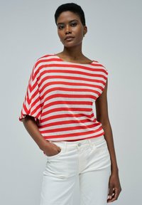 Camiseta a rayas con escote ancho y mangas cortas; rayas rojas y blancas sobre una tela ligera. Se lleva con pantalones blancos, con las manos en los bolsillos.