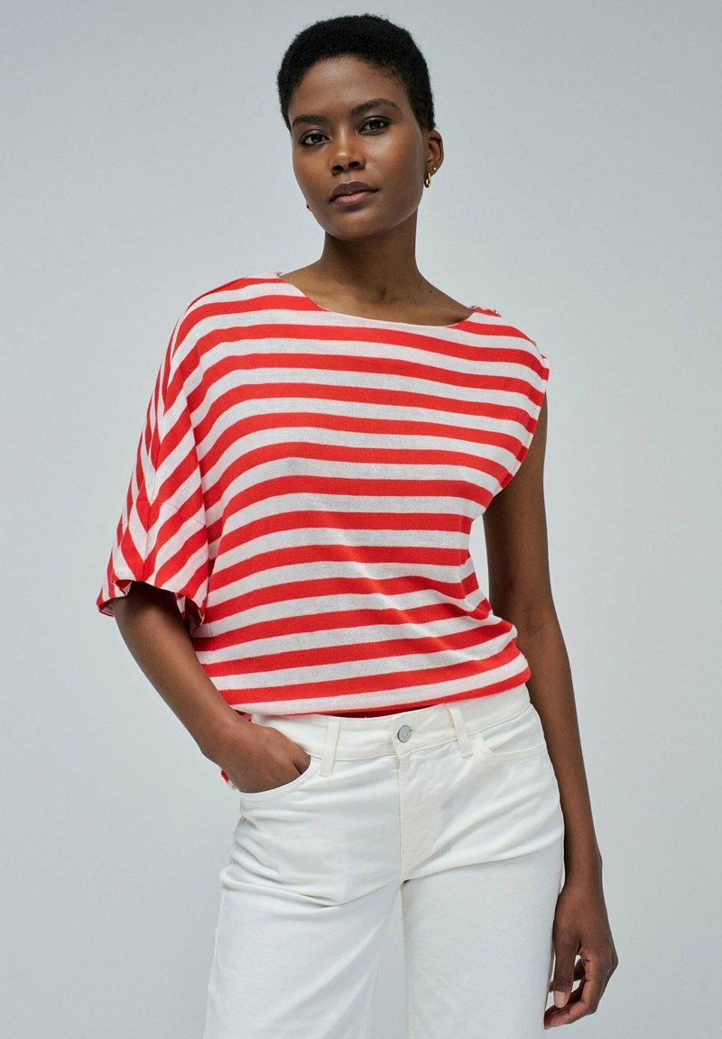 Camiseta a rayas con escote ancho y mangas cortas; rayas rojas y blancas sobre una tela ligera. Se lleva con pantalones blancos, con las manos en los bolsillos.