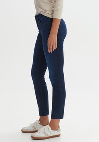 Schmal geschnittene, dunkelblaue Jeans aus Denim mit glatter Textur, über mid-rise Bund und dezenten Gesäßtaschen, kombiniert mit weißen Sneakers.