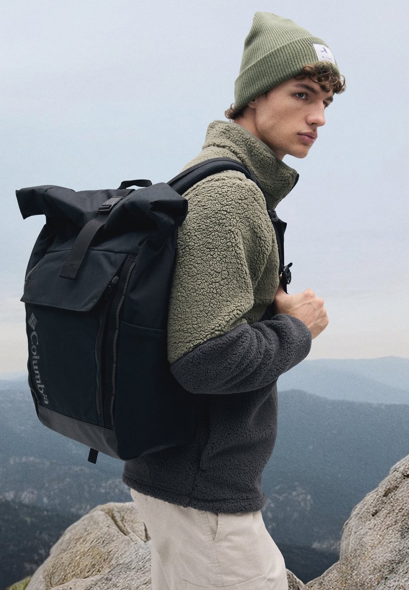 Black roll-top backpack with adjustable straps, gray fleece jacket, and olive green beanie; outdoors with rocky terrain and mountains in background.