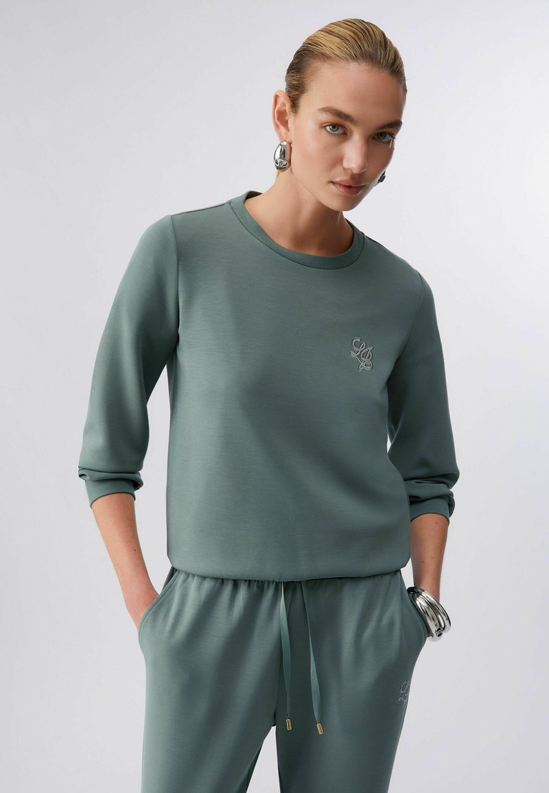 Sweat-shirt vert avec encolure ronde et manches trois-quarts, présentant un logo brodé et une texture lisse et extensible.
