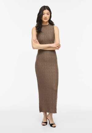 Kobieta o długich ciemnych włosach, ubrana w brązową, bezrękawną, teksturowaną suknię maxi oraz czarne sandały na obcasie z odkrytymi palcami, stojąca ze skrzyżowanymi rękami.
