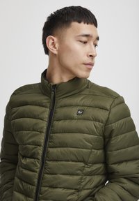 Jeune homme portant une veste matelassée verte zippée, regardant de côté sur un fond clair uni.