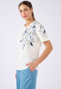 Mujer con cabello castaño que lleva una camisa blanca de manga corta con patrones de hojas azules y rosas y pantalones azul claro, con la mano en el bolsillo.