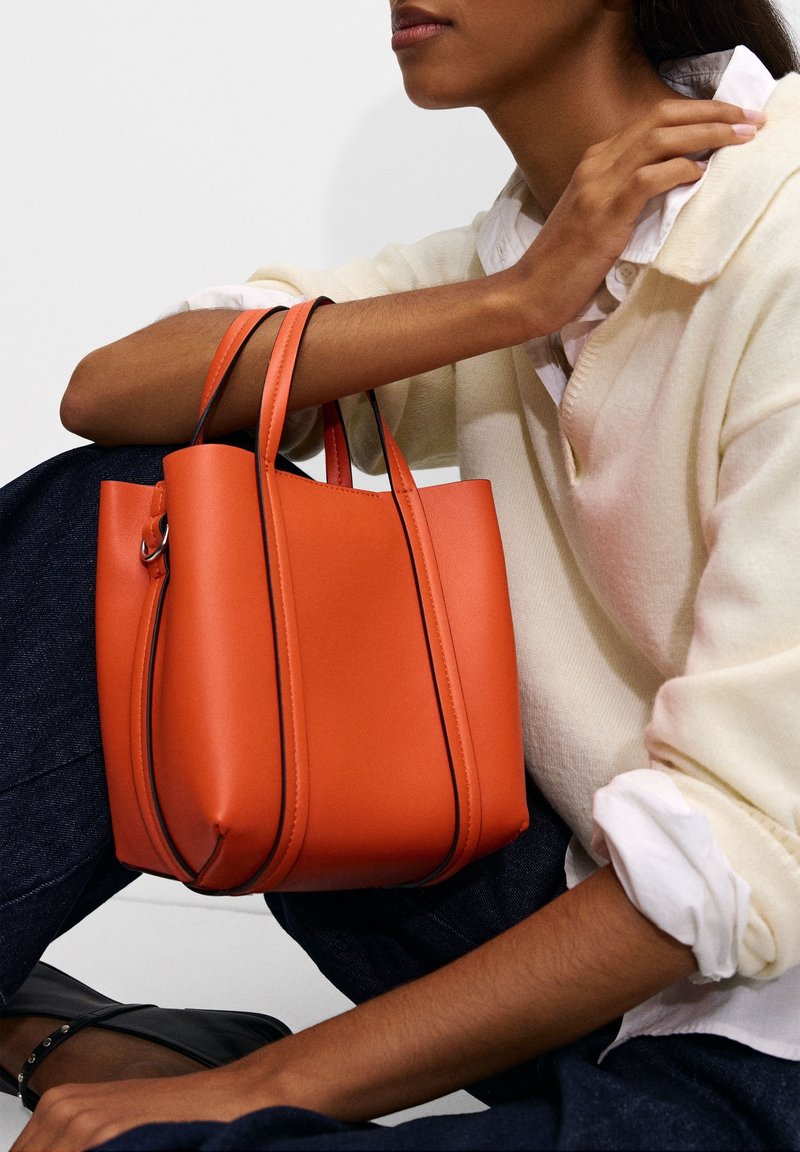 Femme portant un pull crème et un jean foncé tenant un sac à main en cuir orange assise avec un genou relevé sur fond blanc.