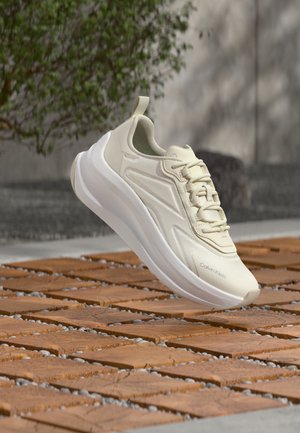 Baskets Calvin Klein beige clair avec une semelle blanche épaisse, flottant au-dessus d'une surface en bois à motif, avec des feuilles vertes floues en arrière-plan.