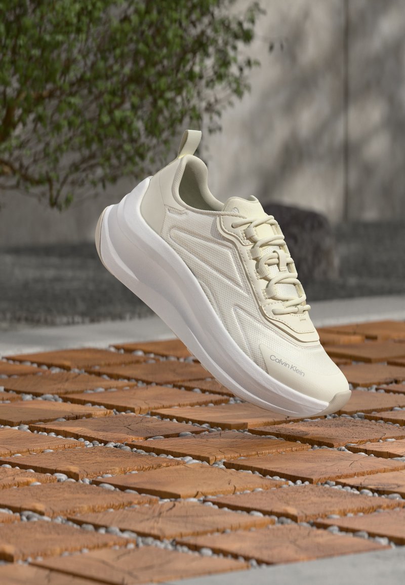 Ανοιχτό μπεζ αθλητικό παπούτσι Calvin Klein με παχιά λευκή σόλα, αιωρούμενο πάνω από μια ξύλινη επιφάνεια με σχέδια, με θολά πράσινα φύλλα στο φόντο.