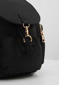Czarny nylonowy plecak z zaokrągloną klapą, złotymi okuciami, zamknięciem na sznurek i boczną kieszenią na zamek. Duża przednia kieszeń z przeszyciami jako ozdobą.