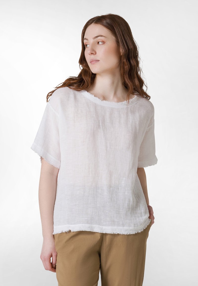 Blusa bianca in lino con maniche corte, scollatura rotonda e bordi sfrangiati. Indossata con pantaloni color khaki. Tessuto morbido e vestibilità ampia.