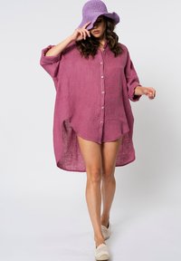 Camicia oversized in lino viola con bottoni frontali, maniche arrotolate e orlo asimmetrico. Abbinata a un cappello intrecciato viola coordinato e calzature leggere.