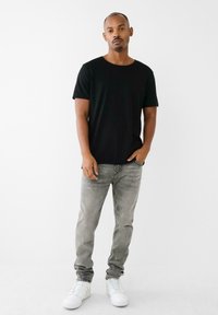 Tricou din bumbac negru, cu mâneci scurte, asortat cu blugi gri deschis și adidași albi. Linii curate și croi lejer. Fundal neutru.