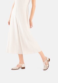 Sandali in pelle beige con tacco chiuso, design a strisce e tacco basso, abbinati a una tuta a gamba larga di colore chiaro.