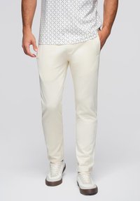 Cremfarbige Hosen mit glatter Textur, ausgestattet mit Seitentaschen und slim fit. Kombiniert mit weißen Sneakers und einem gemusterten Hemd.