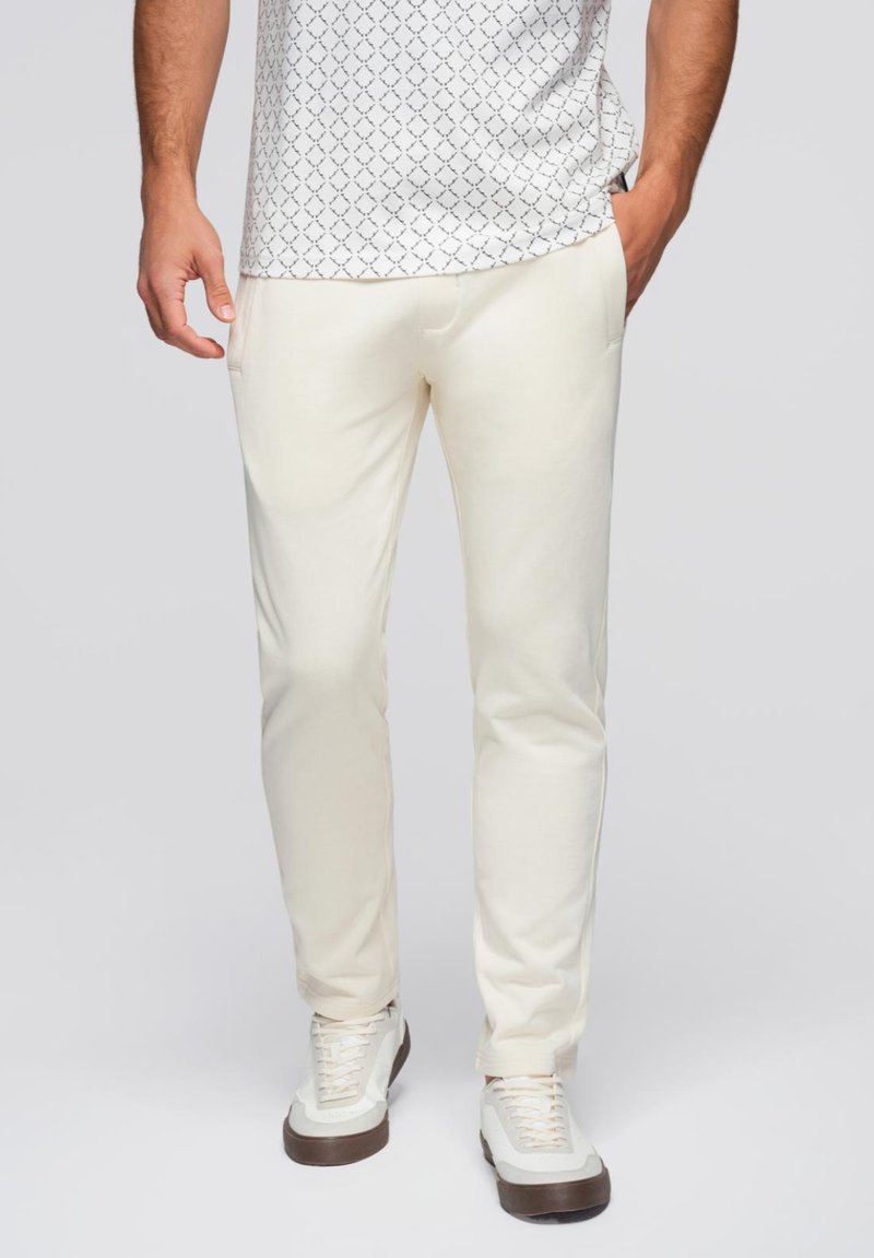 Cremfarbige Hosen mit glatter Textur, ausgestattet mit Seitentaschen und slim fit. Kombiniert mit weißen Sneakers und einem gemusterten Hemd.