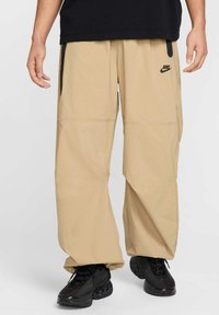 Pantalons cargo beiges avec coupe ample, plusieurs poches et un détail de logo noir, associés à des baskets noires, mettant en avant un matériau texturé.