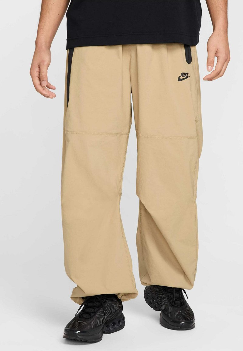 Pantalons cargo beiges avec coupe ample, plusieurs poches et un détail de logo noir, associés à des baskets noires, mettant en avant un matériau texturé.