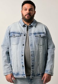 Chaqueta de mezclilla azul clara con cierre de botones, dos bolsillos en el pecho y un corte relajado, que presenta acentos desgastados y un diseño de cuello clásico.