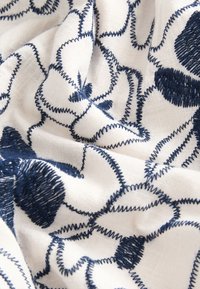 Leinenstoff mit marineblau bestickten Blumenmustern auf weißem Hintergrund, mit strukturierter Oberfläche und sichtbaren Nahtdetails.