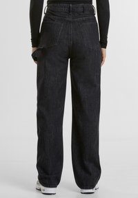 Pantaloni a gamba larga in denim nero con vita alta, dotati di tasche posteriori e una finitura testurizzata. Indossati con sneakers bianche.
