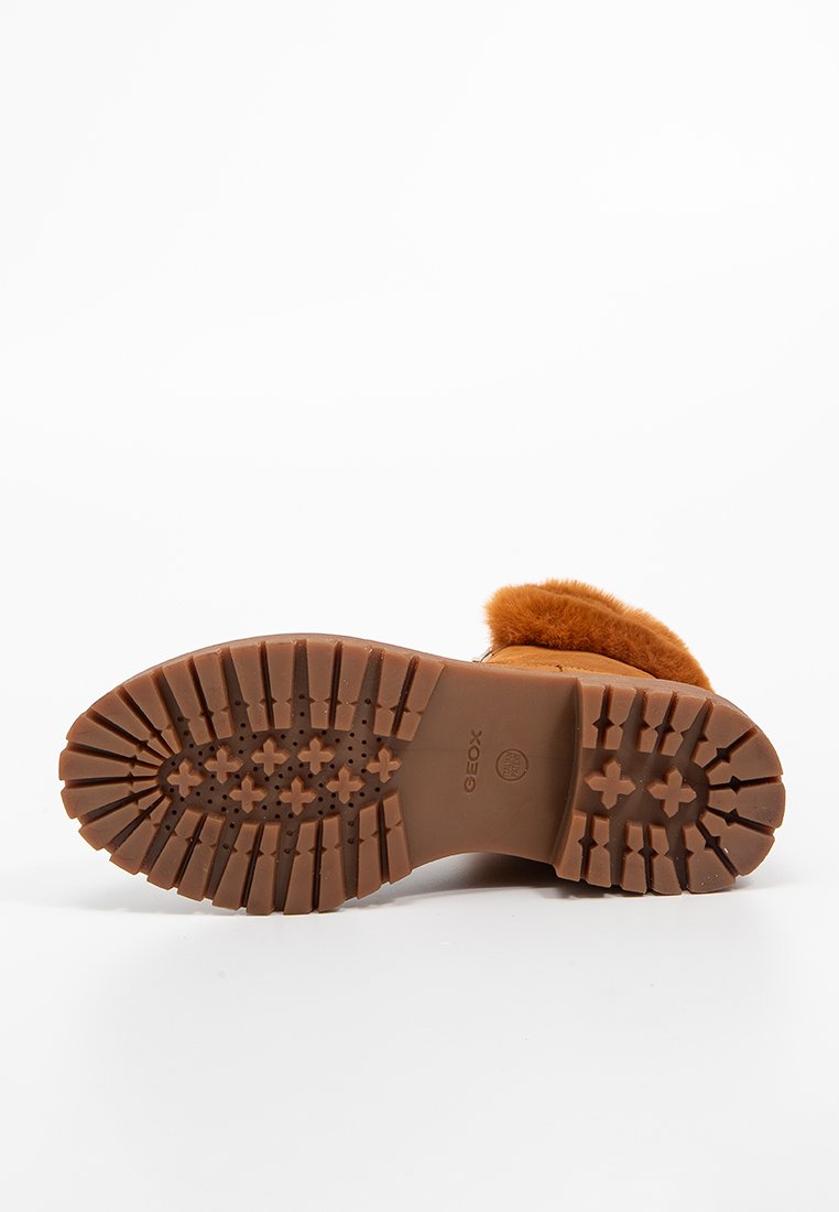 Bota marrón con suela de goma texturizada, forro de piel sintética en la parte superior y forma redondeada. Presenta profundas ranuras para tracción.