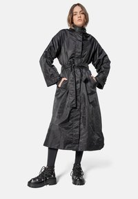 Trench coat negro largo hecho de un tejido brillante, con correa a la cintura, mangas oversize y dos bolsillos frontales. Combinado con botas gruesas negras con cordones.