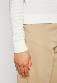 Main droite posée contre un pantalon beige, portant un haut blanc texturé à manches longues avec un poignet côtelé.