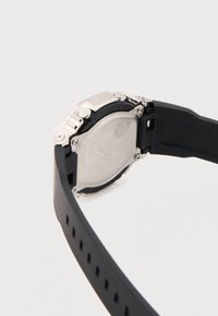 Mustast silikoonist rihmaga käekell hõbedase roostevabast terasest korpusega, millel on mustriline metallist tagakülg ja reguleeritava pandlaga sulgur.