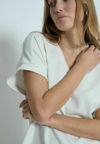 Top bianco a V con maniche corte arrotolate. Tessuto morbido e liscio con una vestibilità rilassata e dettagli di cucitura minimi. Braccia incrociate davanti.
