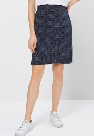 Femme portant une jupe bleue marine à motifs, longueur genou, des baskets blanches et un top blanc sur un fond clair uni.