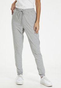 Vrouw in lichtgrijze sweatpants met trekkoord, witte sneakers en een wit T-shirt, staande met één hand in de zak.