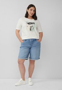 Frau in weißem T-Shirt mit Grafik, hellblauen denim Shorts und weißen Sneakers, die mit den Händen in den Taschen steht.