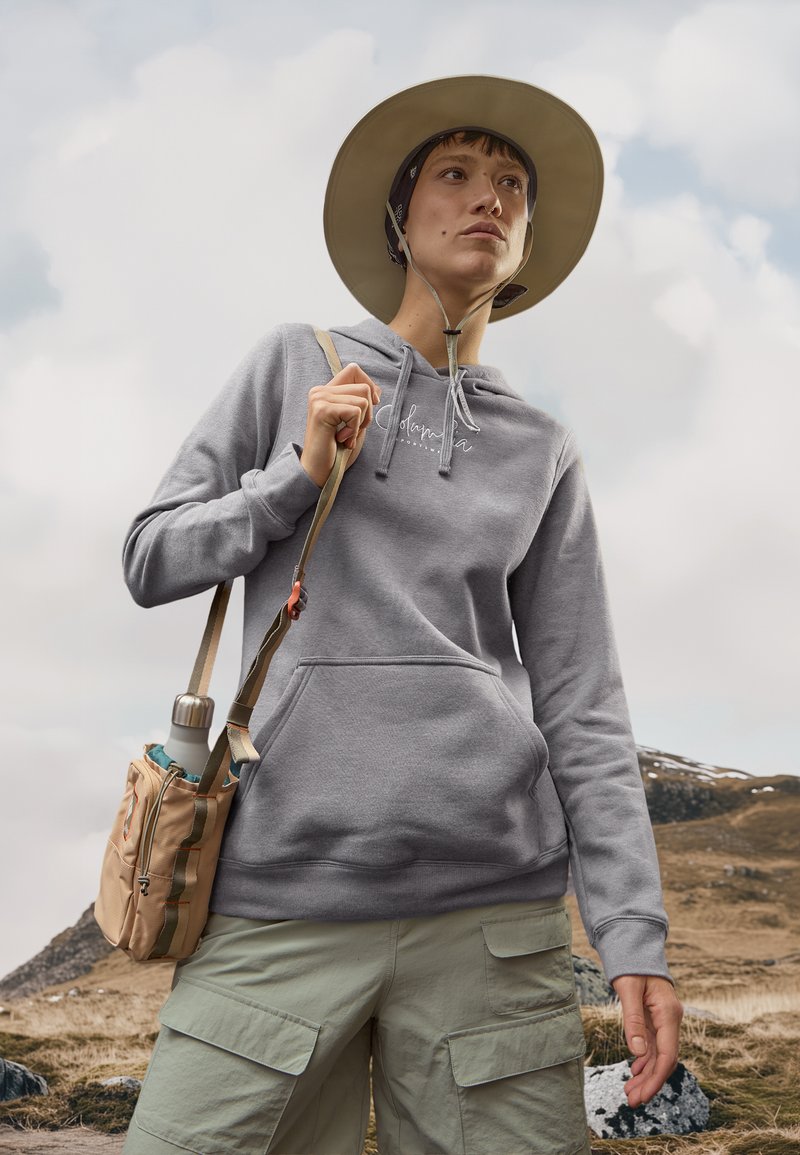 Sudadera de algodón gris con un bolsillo frontal, combinada con pantalones cortos de cargo en verde claro. El sujeto lleva una bolsa beige y usa un sombrero de ala ancha.