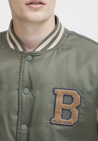 Giacca bomber verde oliva con colletto beige a righe, bottoni a pressione e dettaglio in tessuto marrone a forma di lettera "B" con contorno blu. Texture liscia.
