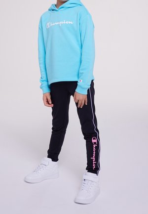 Niño con sudadera con capucha Champion azul claro, pantalones deportivos Champion negros con logo rosa y zapatillas blancas de caña alta, de pie sobre un fondo liso.