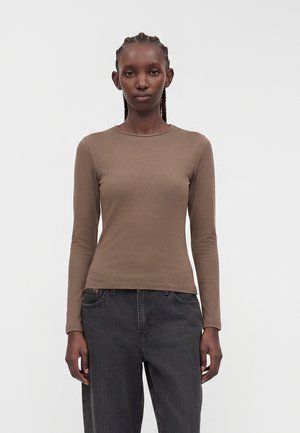 Kvinde med fletninger iført en tætsiddende brun langærmet top og afslappede sorte jeans, stående mod en simpel hvid baggrund.