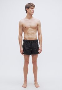 Μαύρο μαγιό σορτς με ελαστική μέση και κορδόνι. Διαθέτει αντίθετο μοτίβο λογότυπου στην αριστερή πλευρά. Ομαλή υφή, μήκος πάνω από το γόνατο.