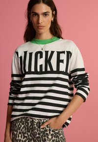 Majica dugih rukava crno-bijelih pruga s velikim natpisom "WICKED". Ima zelenu ovratnik. Uparen s hlačama leopard uzorka. Roza pozadina.