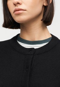 Femme aux cheveux bruns courts portant un cardigan noir boutonné par-dessus un haut rayé à col rond, légèrement tournée vers la gauche.